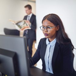 Focused young African American businesswoman working on a computer at her office desk with a colleague in the background